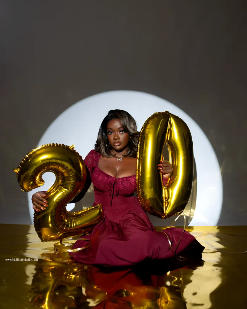 Woman in maroon dress facing camera with both hands gripping the gold number-20 balloon digits, mirror floor