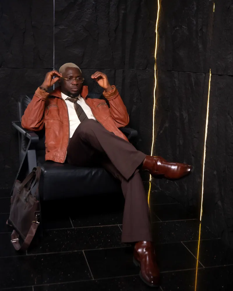 Full body seated in tan leather jacket, black tile floor, neon light accent