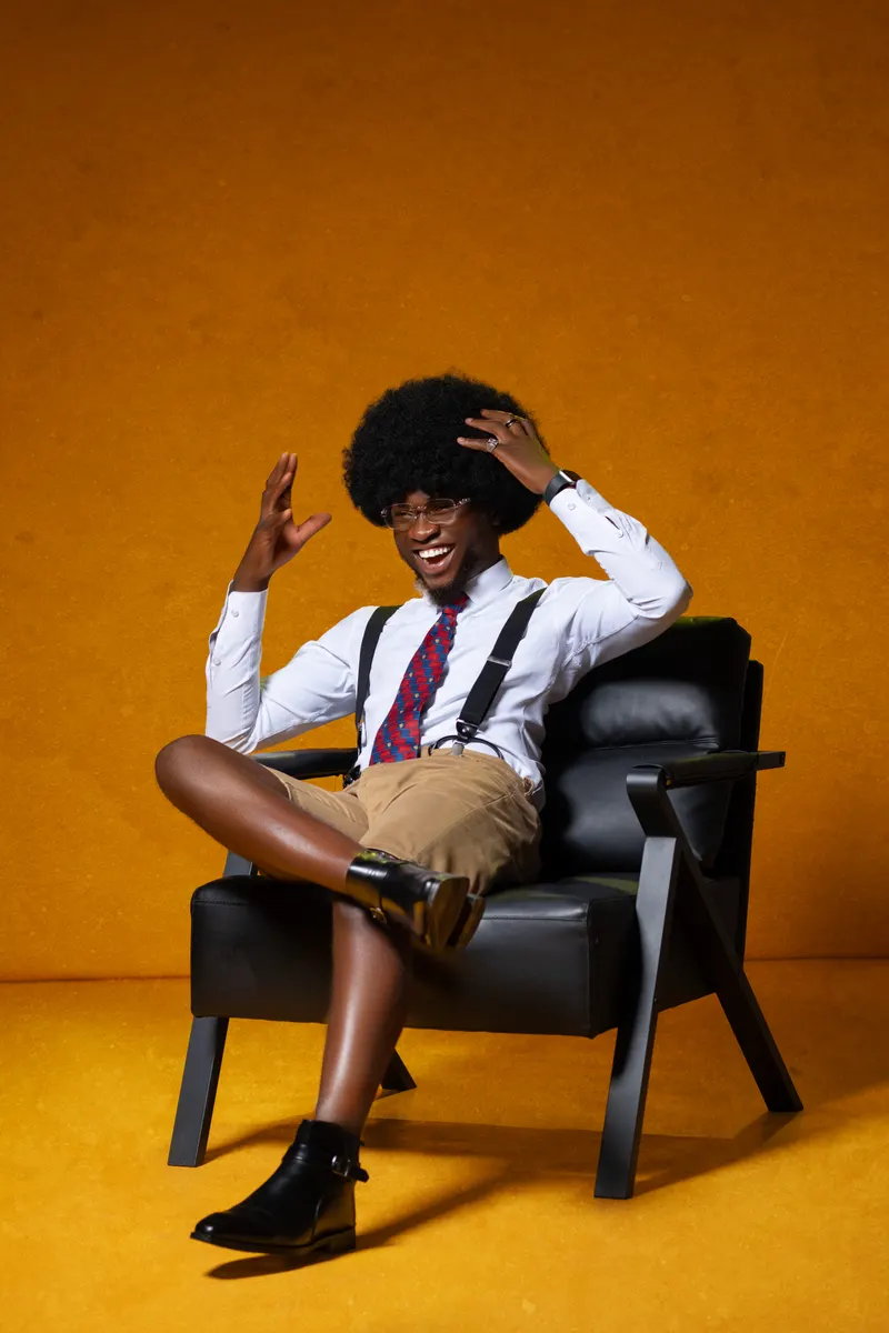 Same afro character, playful seated pose in suspenders/shorts, orange backdrop
