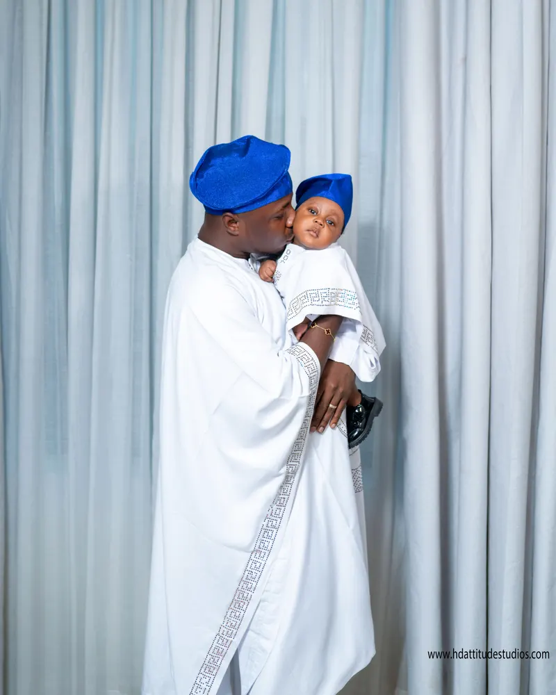Father in white agbada and blue fila kissing baby's cheek, baby in matching blue cap, blue curtain backdrop