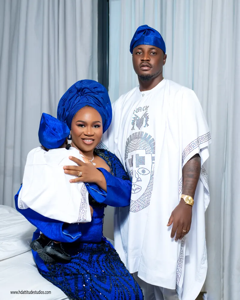Parents in matching royal-blue aso-oke with baby in blue cap, white curtain backdrop