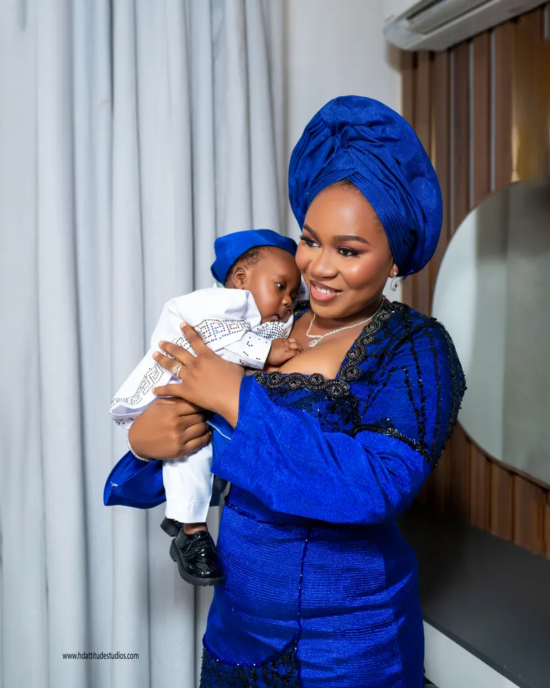 Mother in blue and black beaded gele outfit cradling baby in blue cap and white sequin outfit