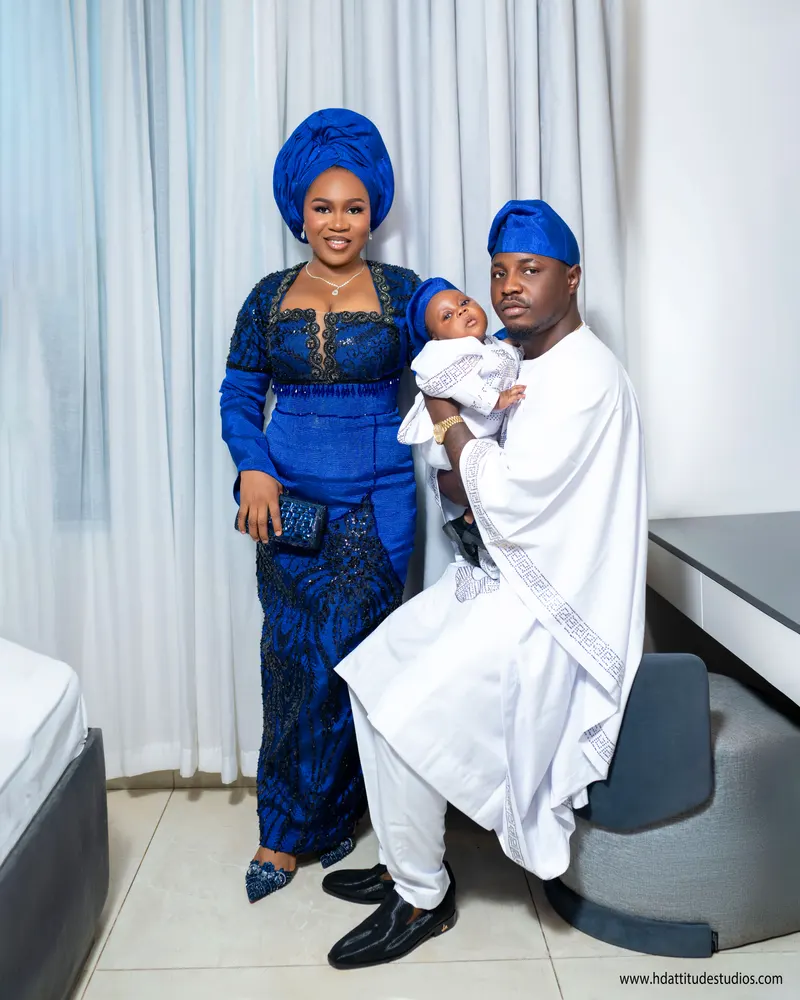 Family trio seated, father in white agbada holding baby, mother in blue standing beside