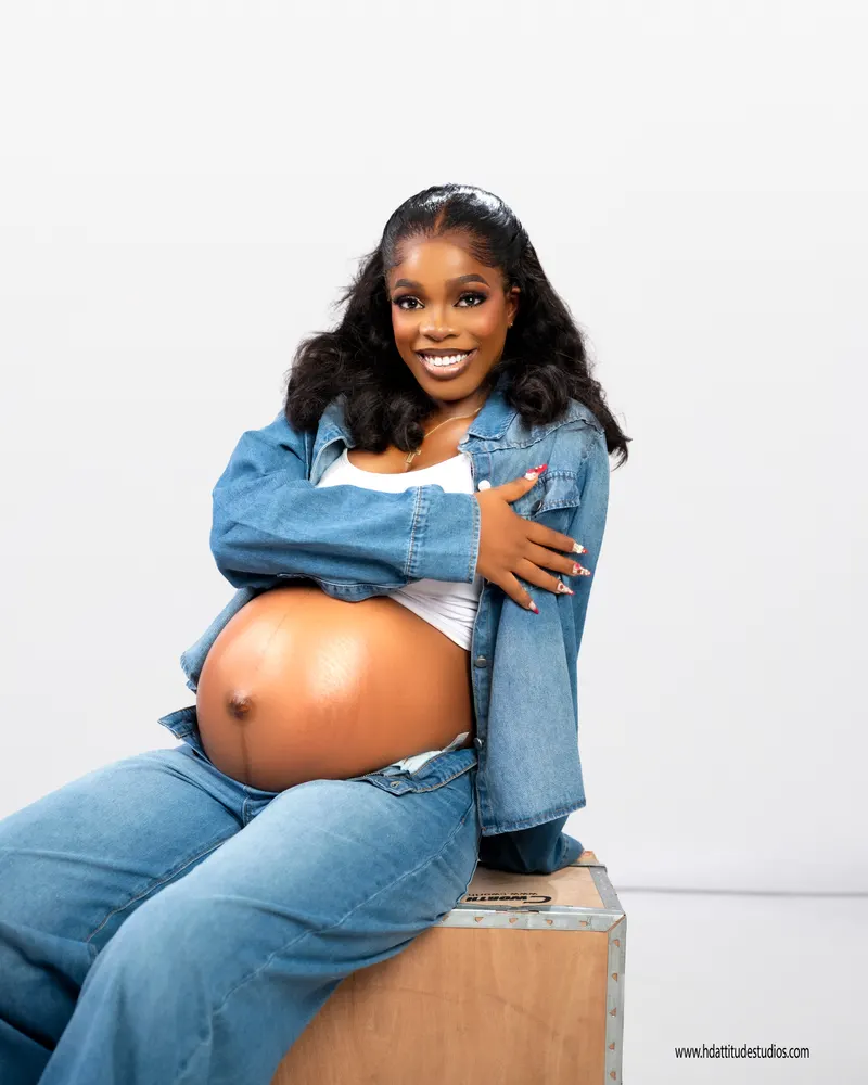 Same denim maternity set, seated on crate, arm draped over bump