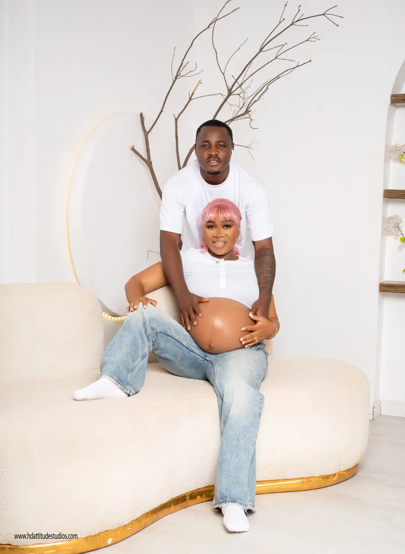 Maternity portrait, full seated pose on sofa, both hands on belly