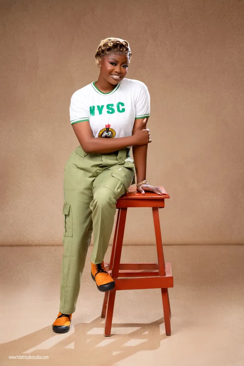 NYSC uniform milestone portrait, seated on stool, tan backdrop