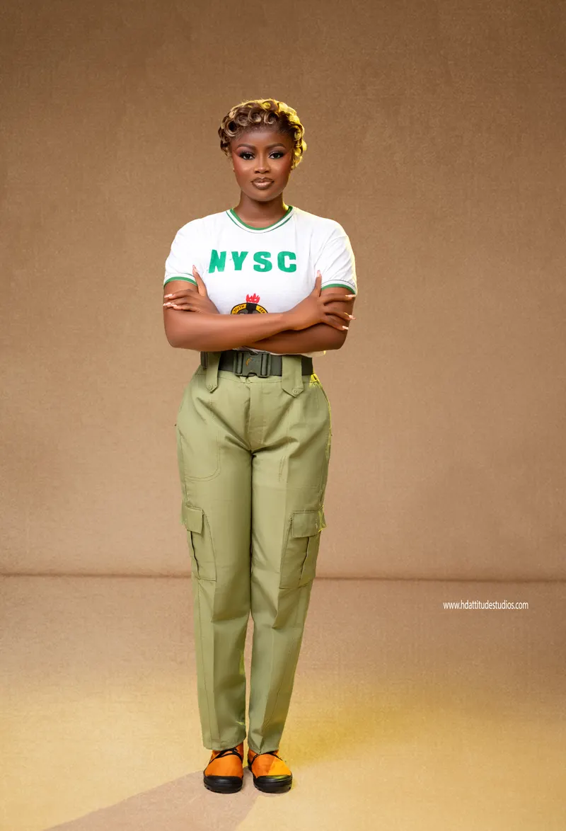 Same NYSC set, standing arms-crossed, tan backdrop