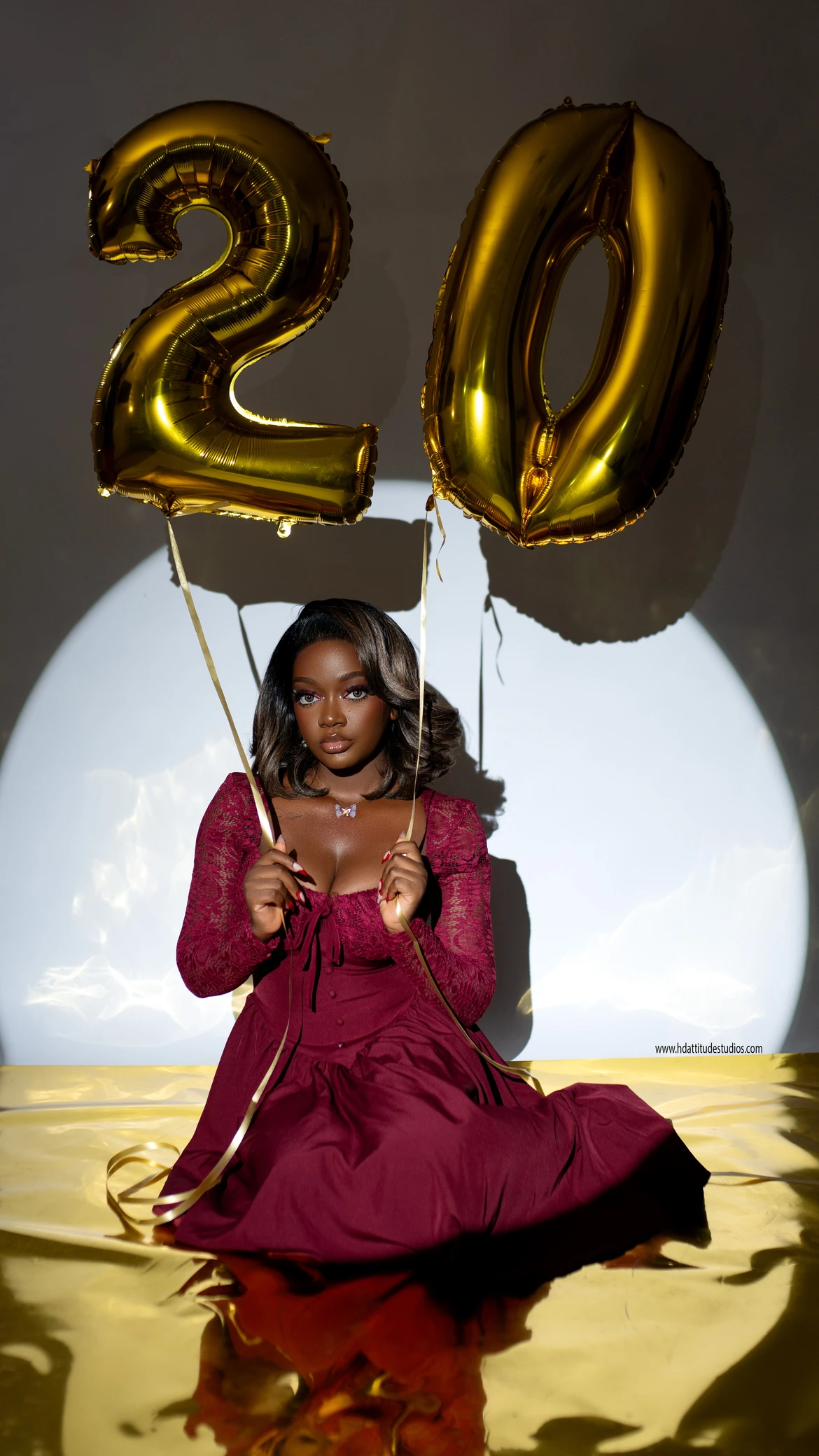 Woman in maroon dress facing camera with both hands gripping the gold number-20 balloon digits, mirror floor