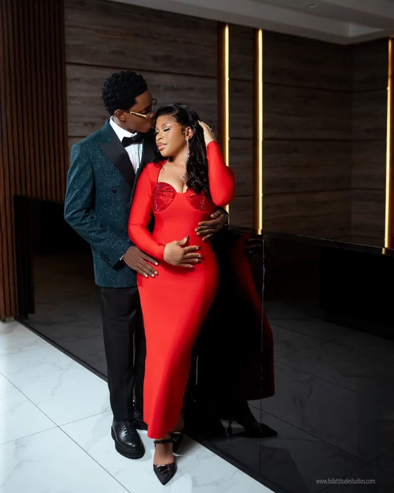 Couple embracing beside black reception desk, man in green glitter blazer kissing woman's hairline