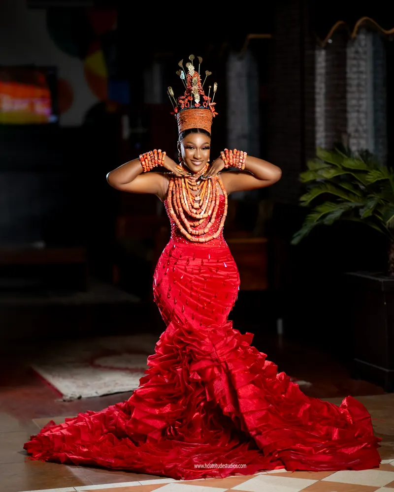 Solo bride in elaborate coral-bead crown and ruffled red mermaid gown, hands raised to headpiece