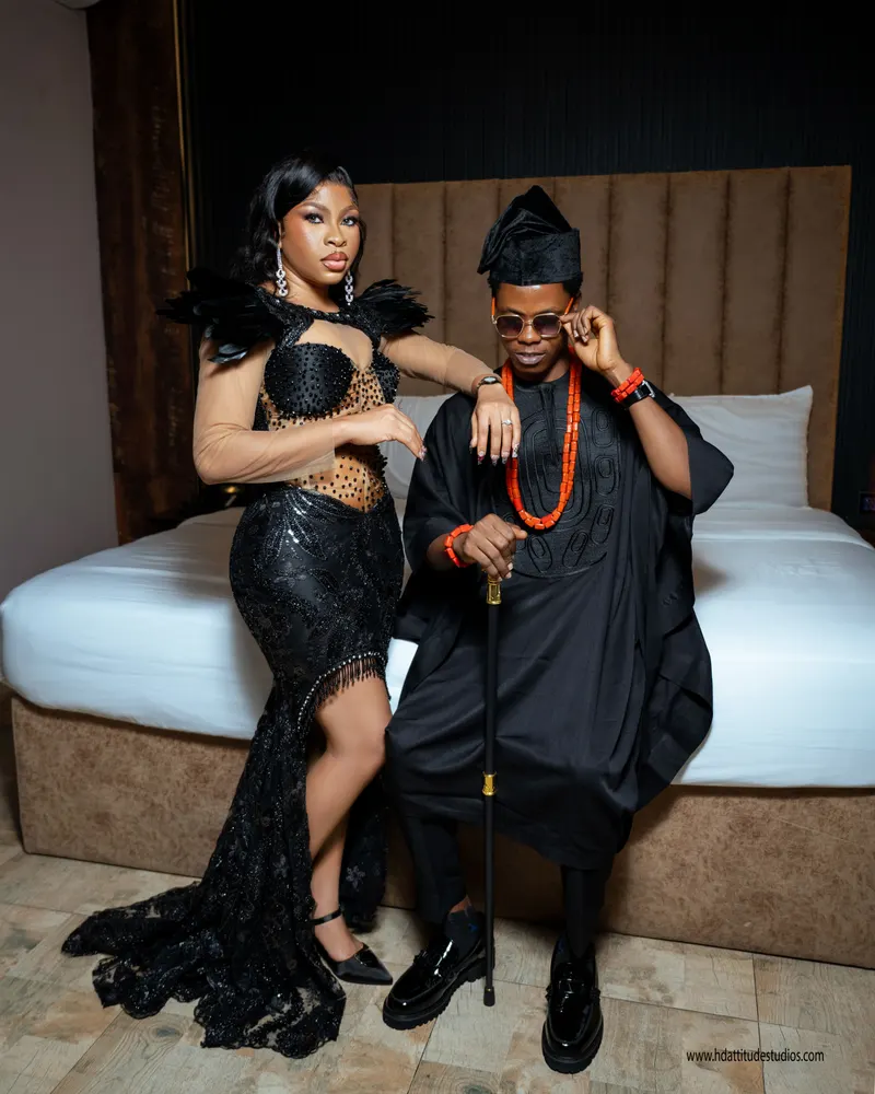 Couple on bed edge, woman in black feather beaded gown, man in black agbada with coral beads and cane