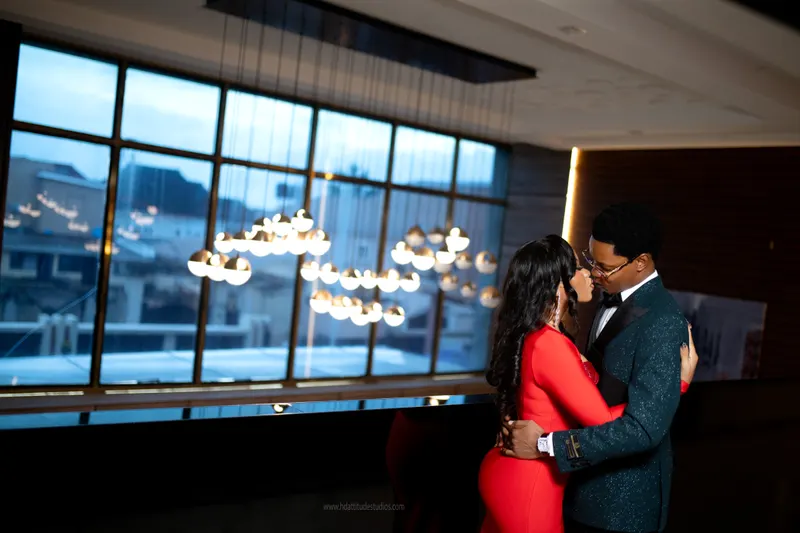 Couple nose to nose in the window and pendant-light lobby, red dress and green glitter tux