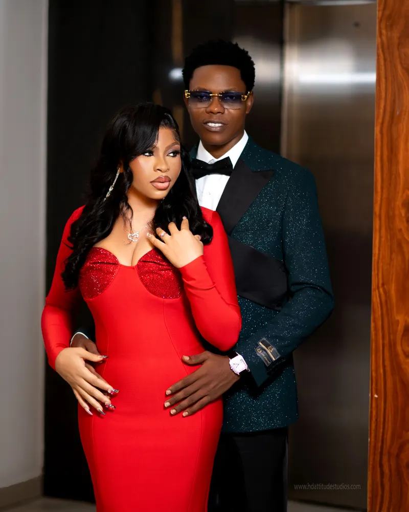 Couple in elevator lobby, woman in red gown, man in sunglasses and green glitter tuxedo